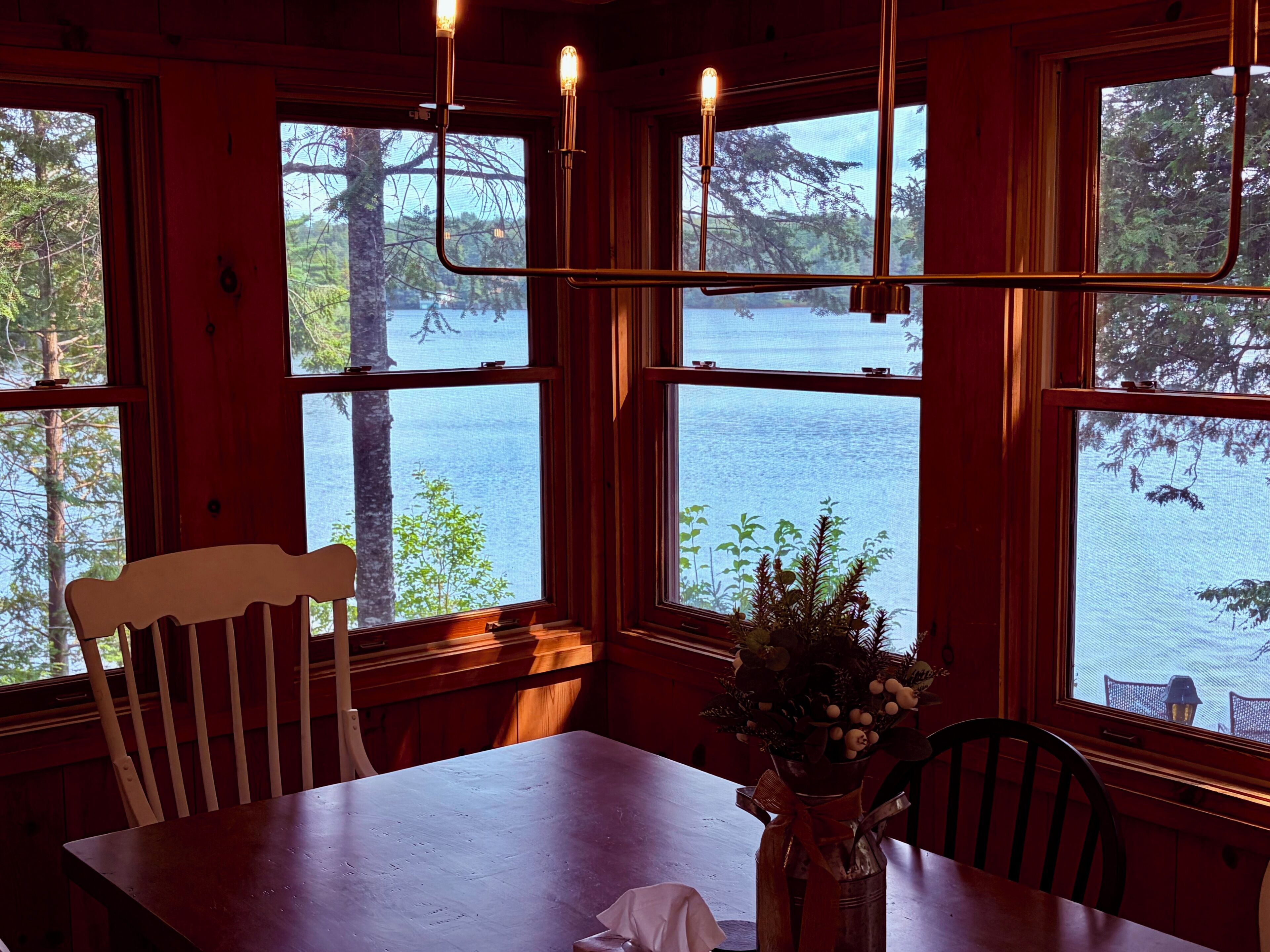 A wooden dining table with chairs, a vase with flowers, and a view of a lake through large windows.