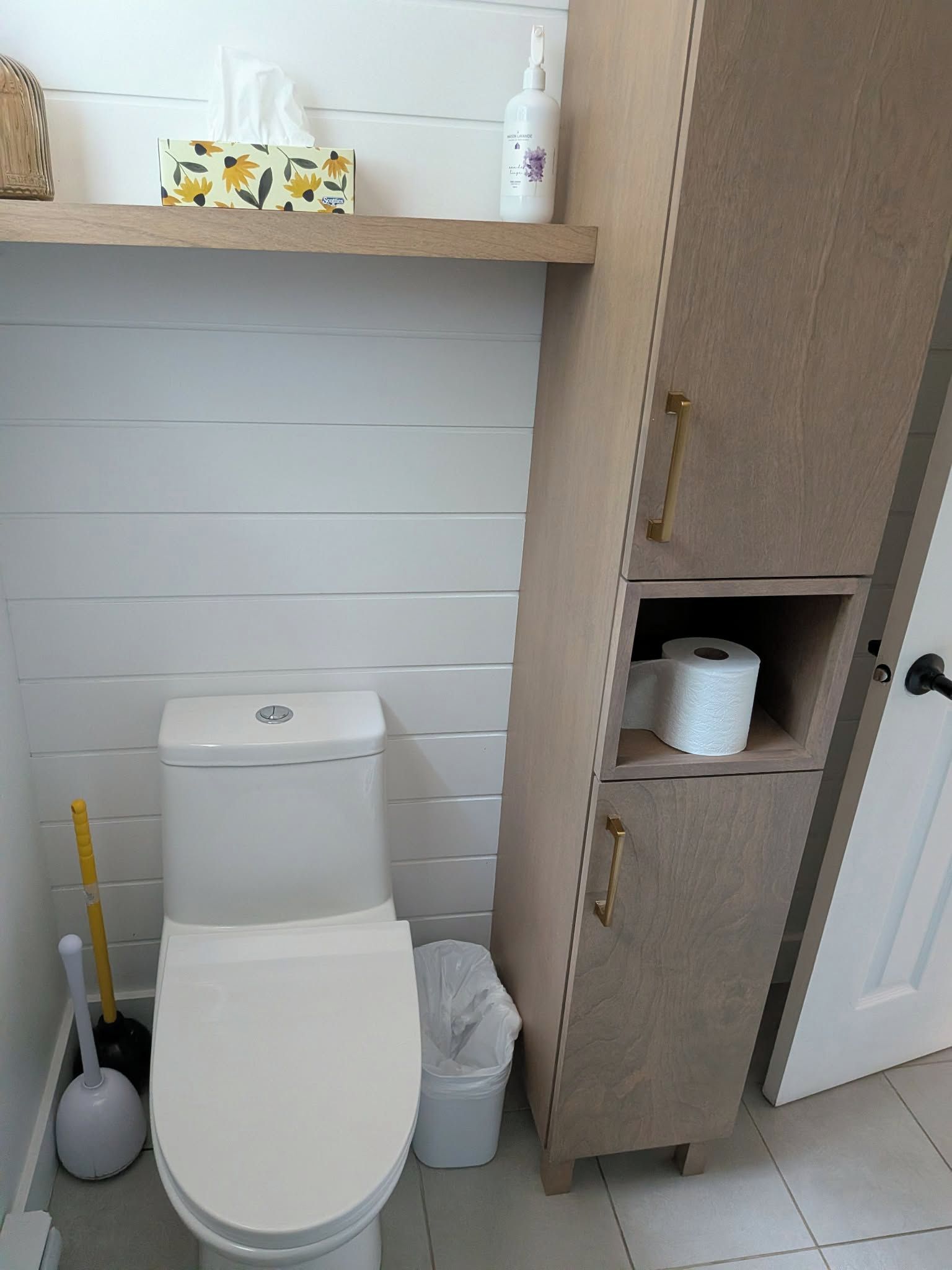 Une salle de bain comprenant des toilettes, un bidet, une poubelle et un meuble en bois avec une étagère au-dessus.