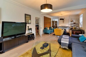 A modern living room with a flat-screen TV, a sofa, a coffee table, and a dining area in the background. - Heritage Holiday Home With Garden (Oxford)