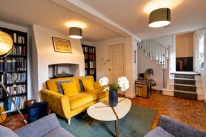 Un salon avec un canapé jaune, une table basse ronde et une bibliothèque remplie de livres. Un escalier mène à un autre niveau.