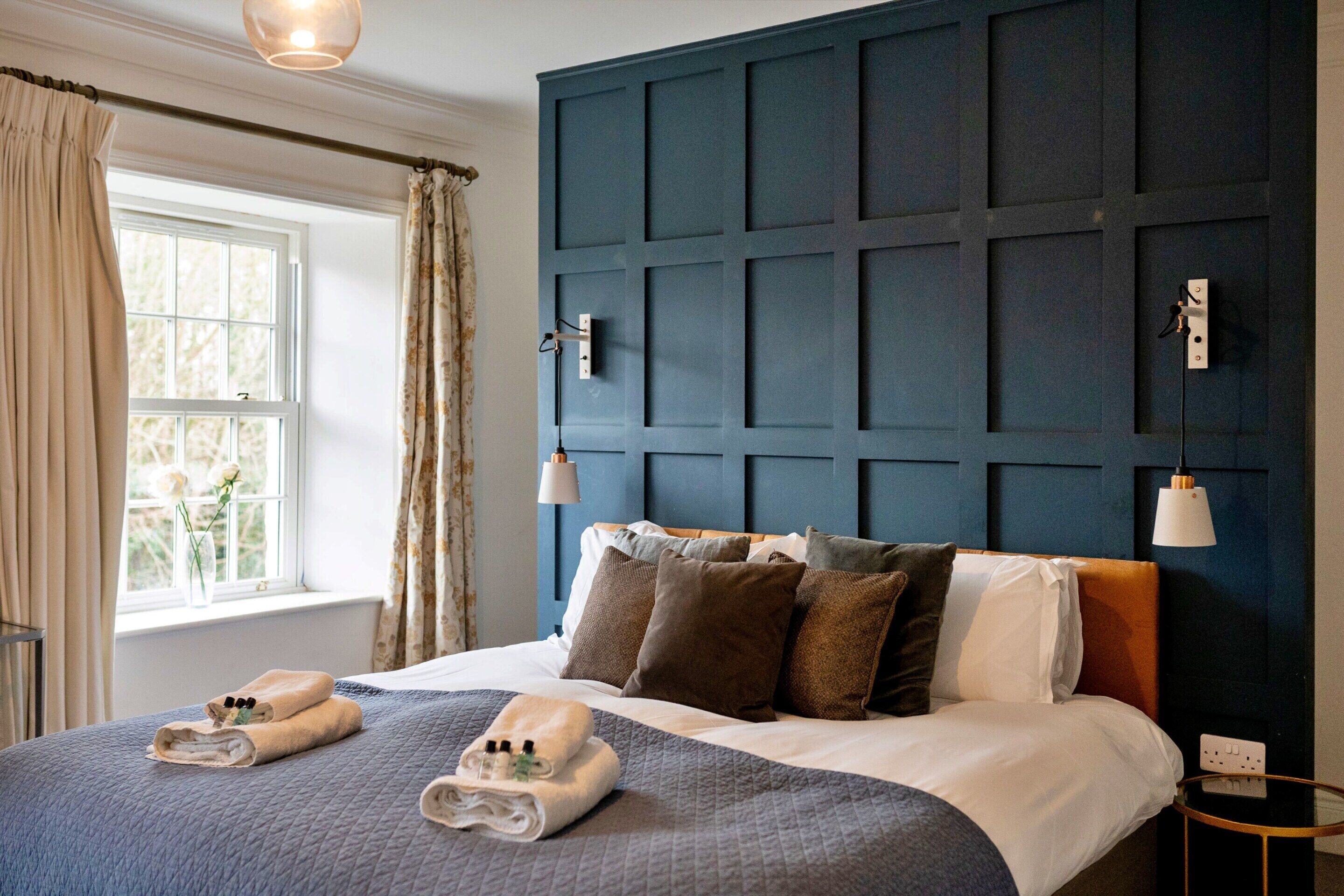 Una cama bien hecha con cabecera azul, una ventana con cortinas y una mesita de noche con lámpara.
