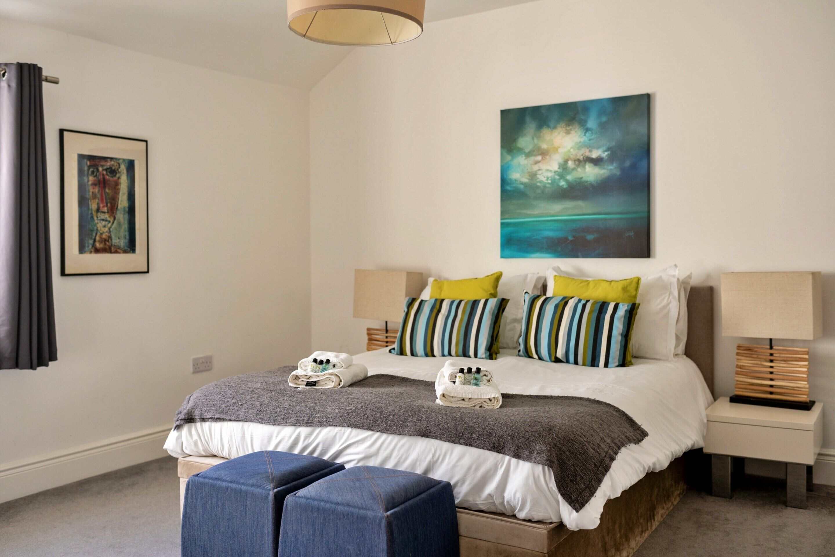 A modern bedroom with a bed, pillows, a nightstand, a lamp, and a painting on the wall.