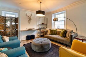 Un salon moderne avec un canapé d’angle, un pouf rond, une table basse et un lampadaire.