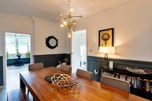 A dining area with a wooden table, chairs, a chandelier, a wall-mounted mirror, and a bookshelf with books. - Heritage Holiday Home With Garden (Oxford)