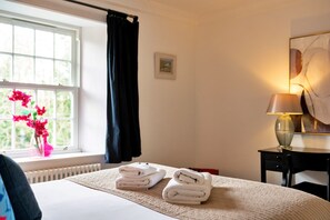 A hotel room with a bed, a window with curtains, a lamp, and a vase with flowers. - Heritage Holiday Home With Garden (Oxford)
