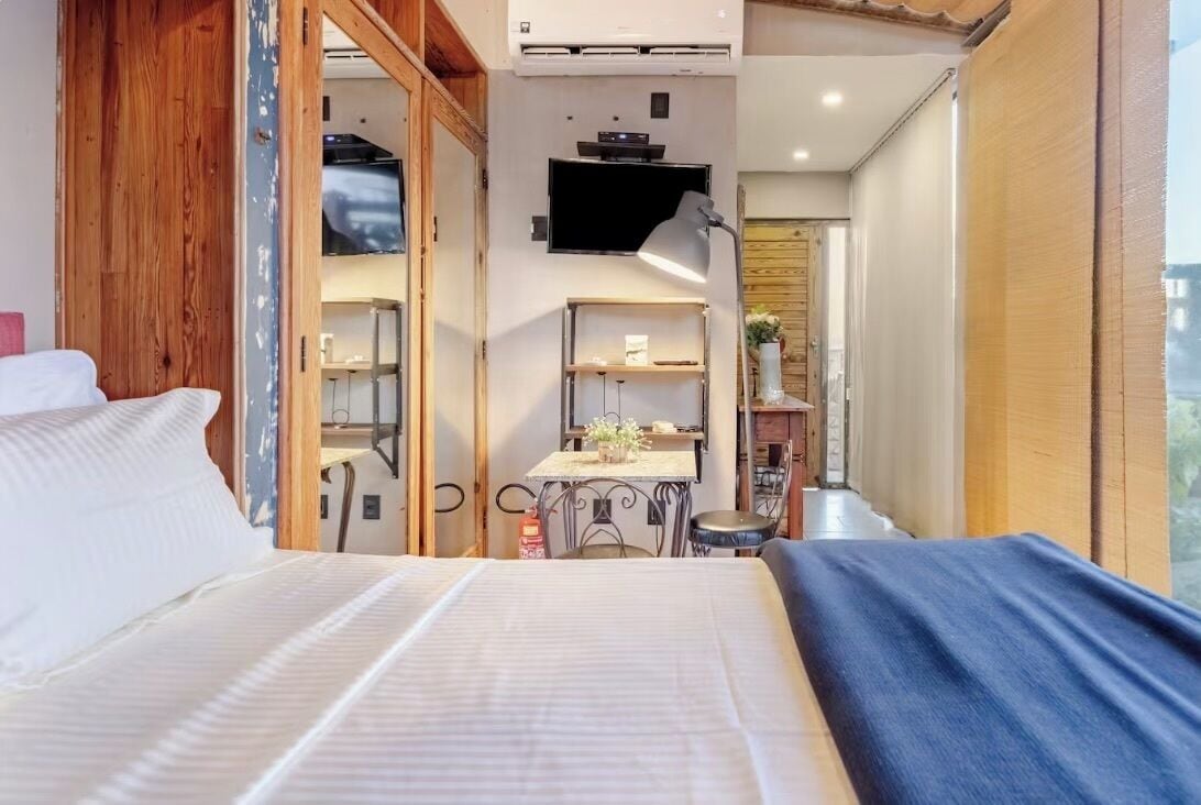 A bedroom with a bed, a desk, and a chair, a television mounted on the wall, and a window.