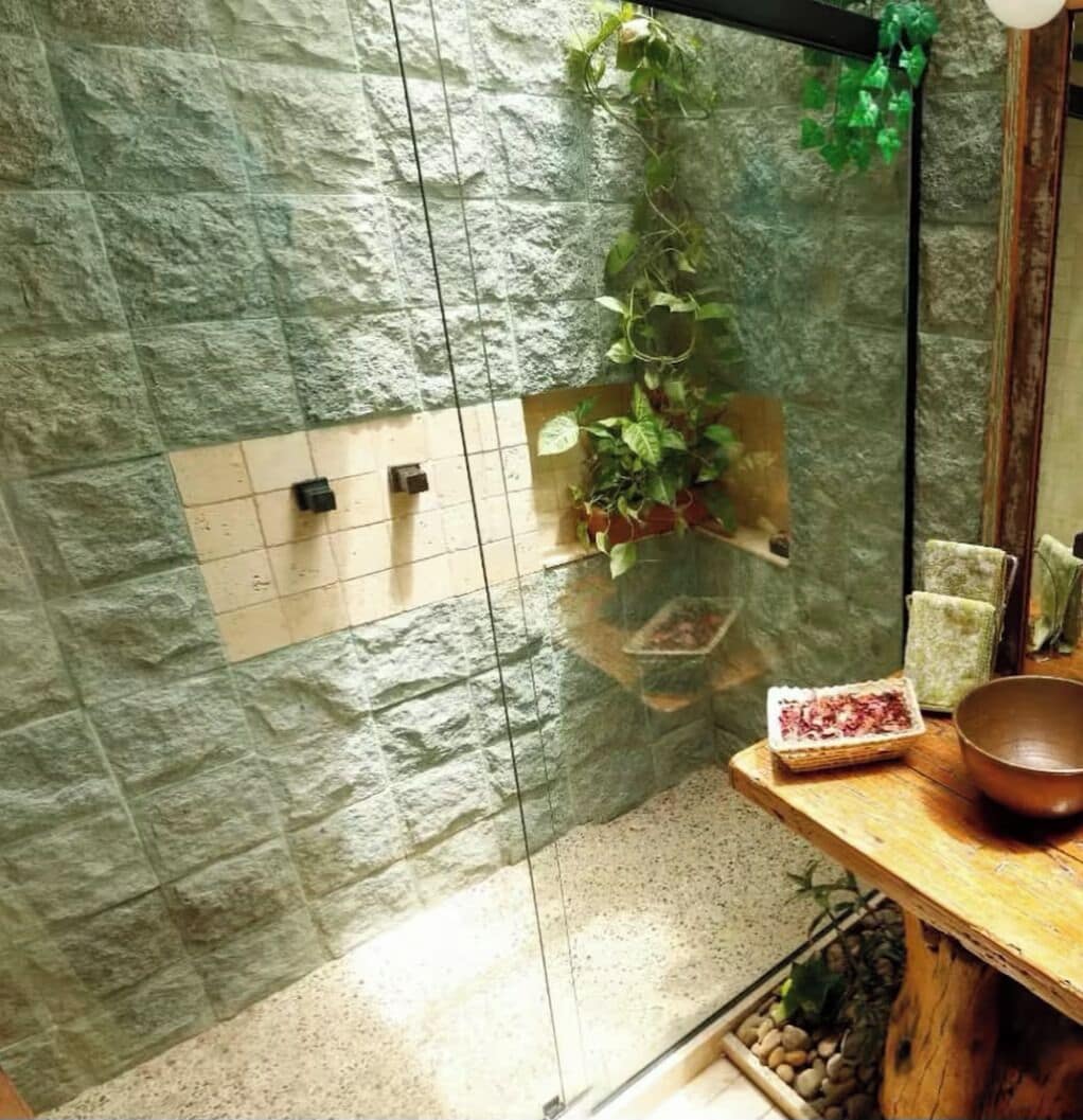 Un baño con pared de piedra, cabina de ducha de vidrio, estante de madera con artículos de aseo y una planta colgante.