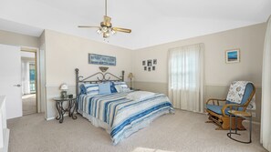 Ein Schlafzimmer mit einem Bett, einem Sessel, einem Deckenventilator, einem Nachttisch und einem Fenster mit Vorhängen.