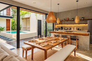 A modern kitchen with a central island, a dining area with a wooden table and benches, and a pool visible through large glass doors. - Remodeled 4BR Boho Villa · Pool Trilogy + Hot Tub (Tulum)