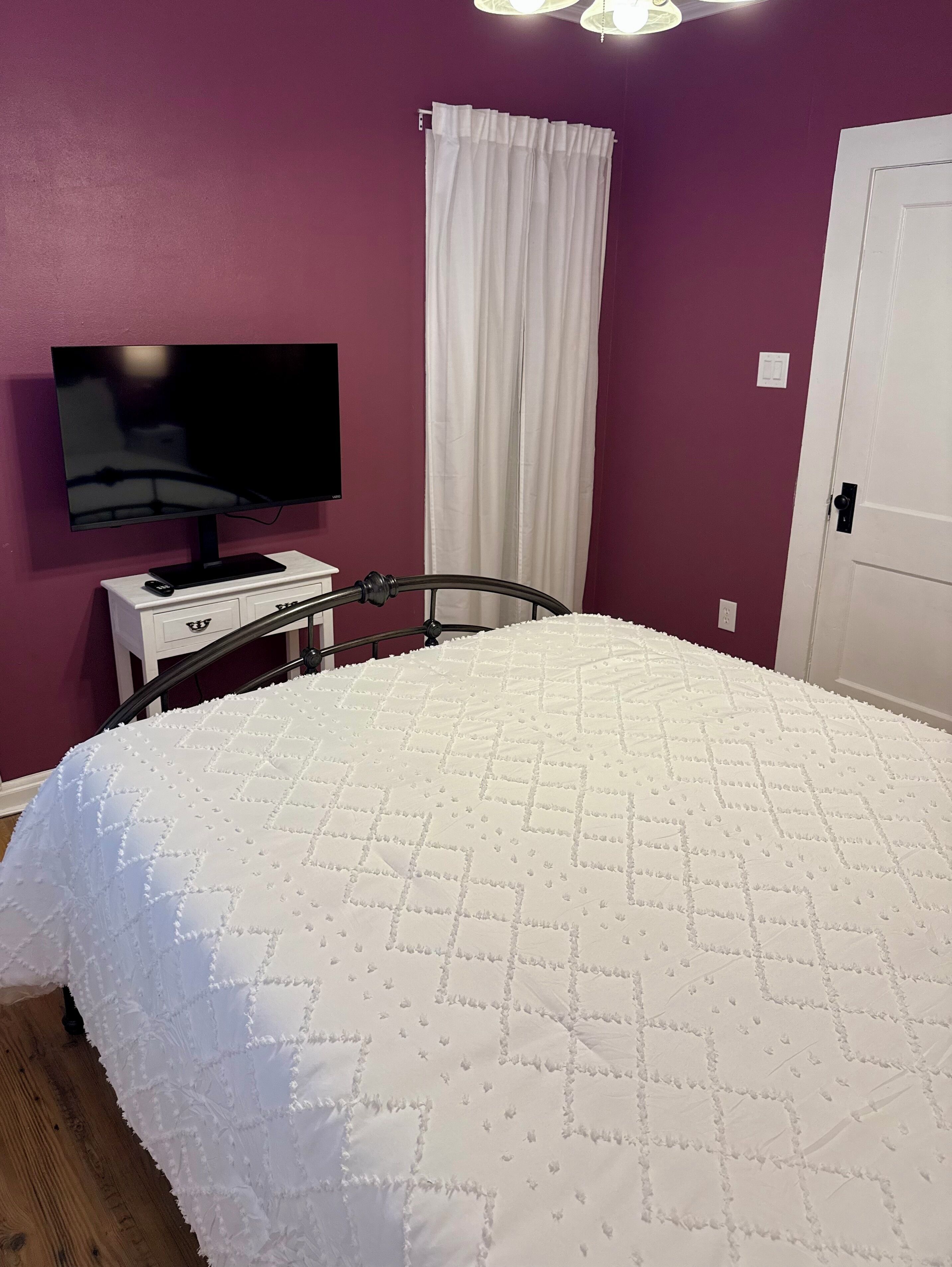 Een slaapkamer met een bed, een televisie op een standaard, een witte deur en een gordijn.