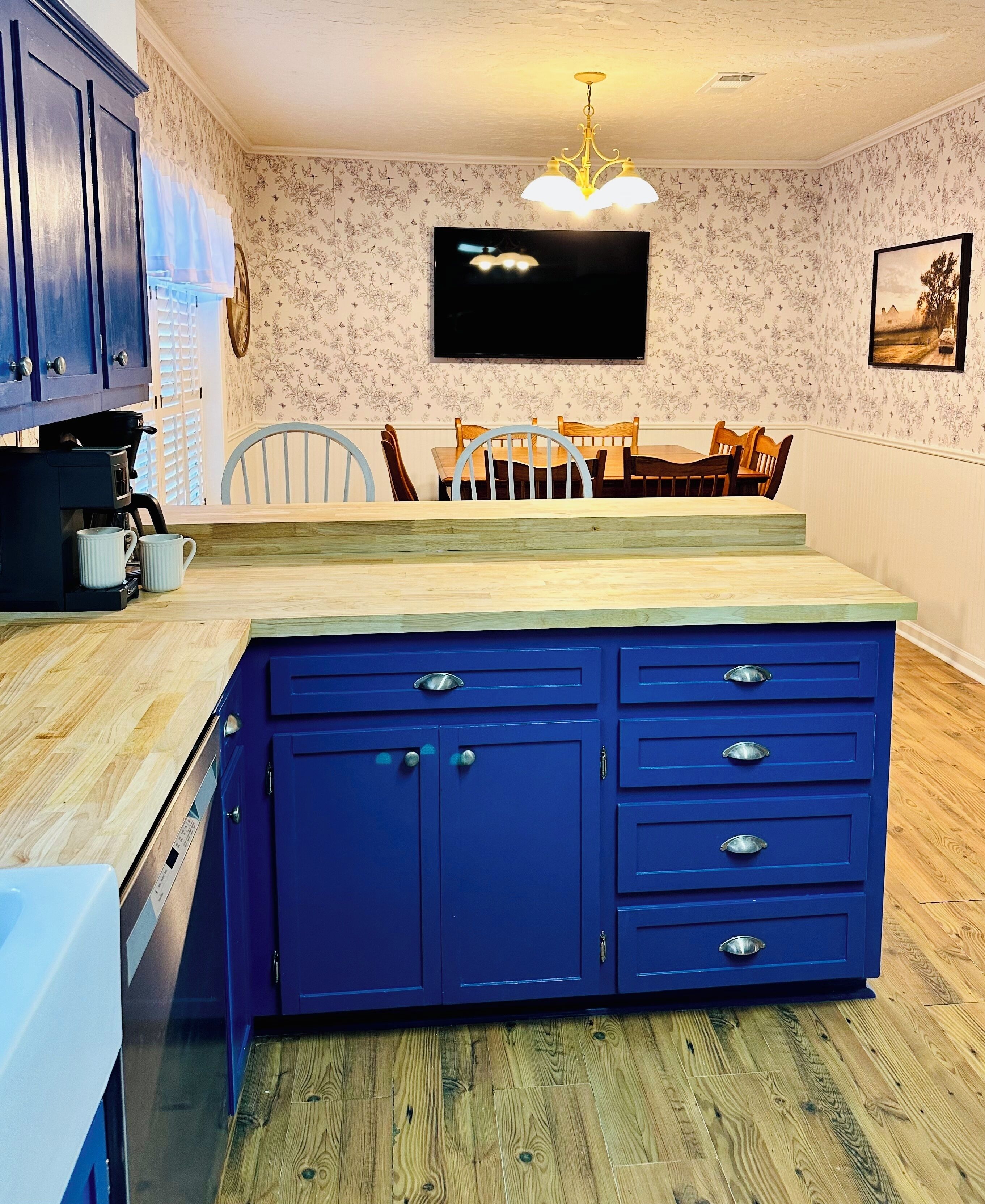 Eine Küche mit einer blauen Insel, Holzschränken, einem Esstisch mit Stühlen, einer Kaffeemaschine und einem an der Wand befestigten Flachbildfernseher.