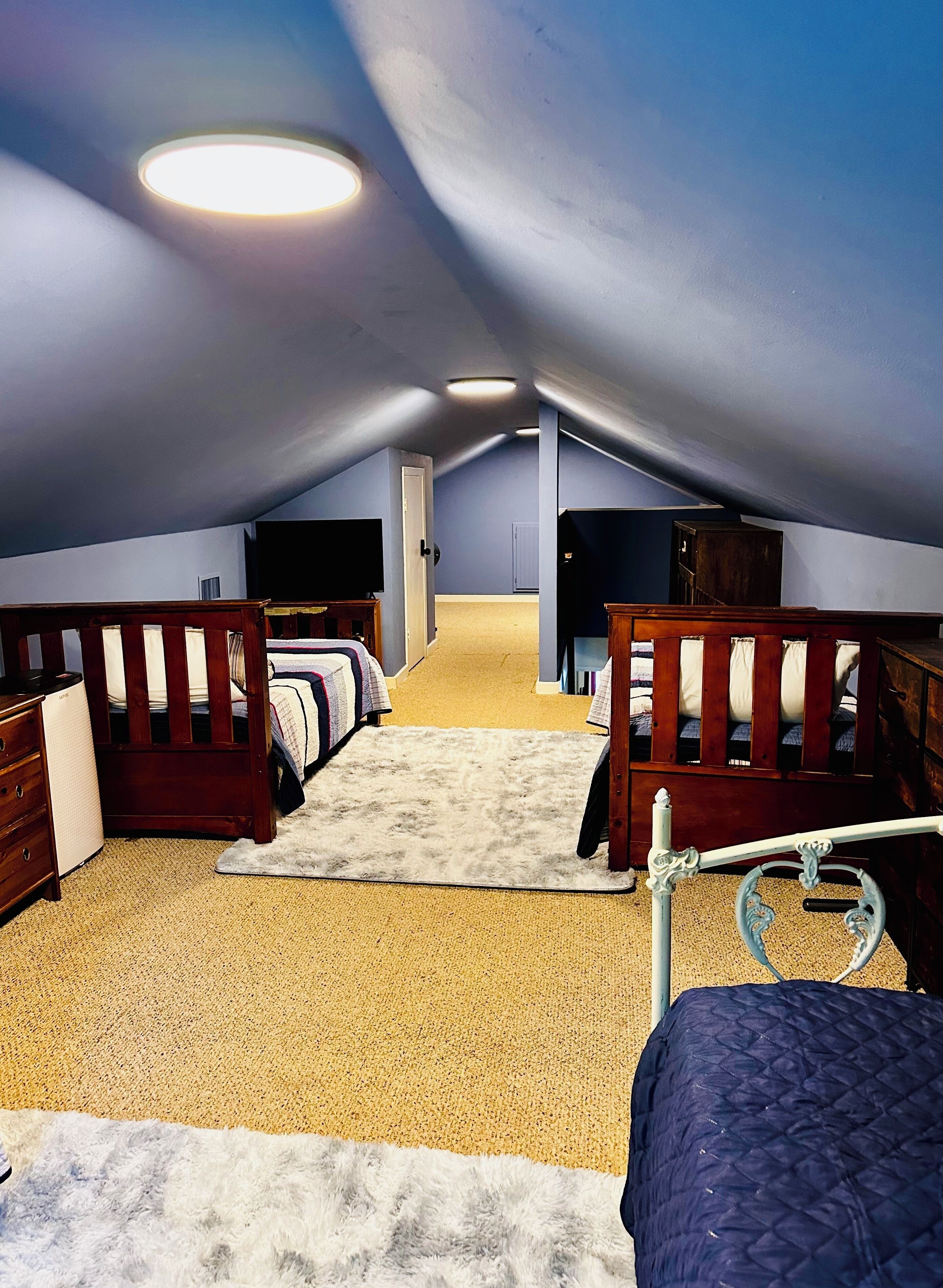 A room with a sloped ceiling, two beds with wooden headboards, a small white dresser, and a television on the wall.