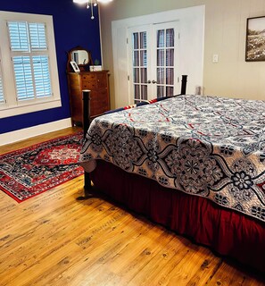 Een slaapkamer met een groot bed, een houten dressoir, een vloerkleed met rood en zwarte patronen, en een deur voorzien van gordijnen.