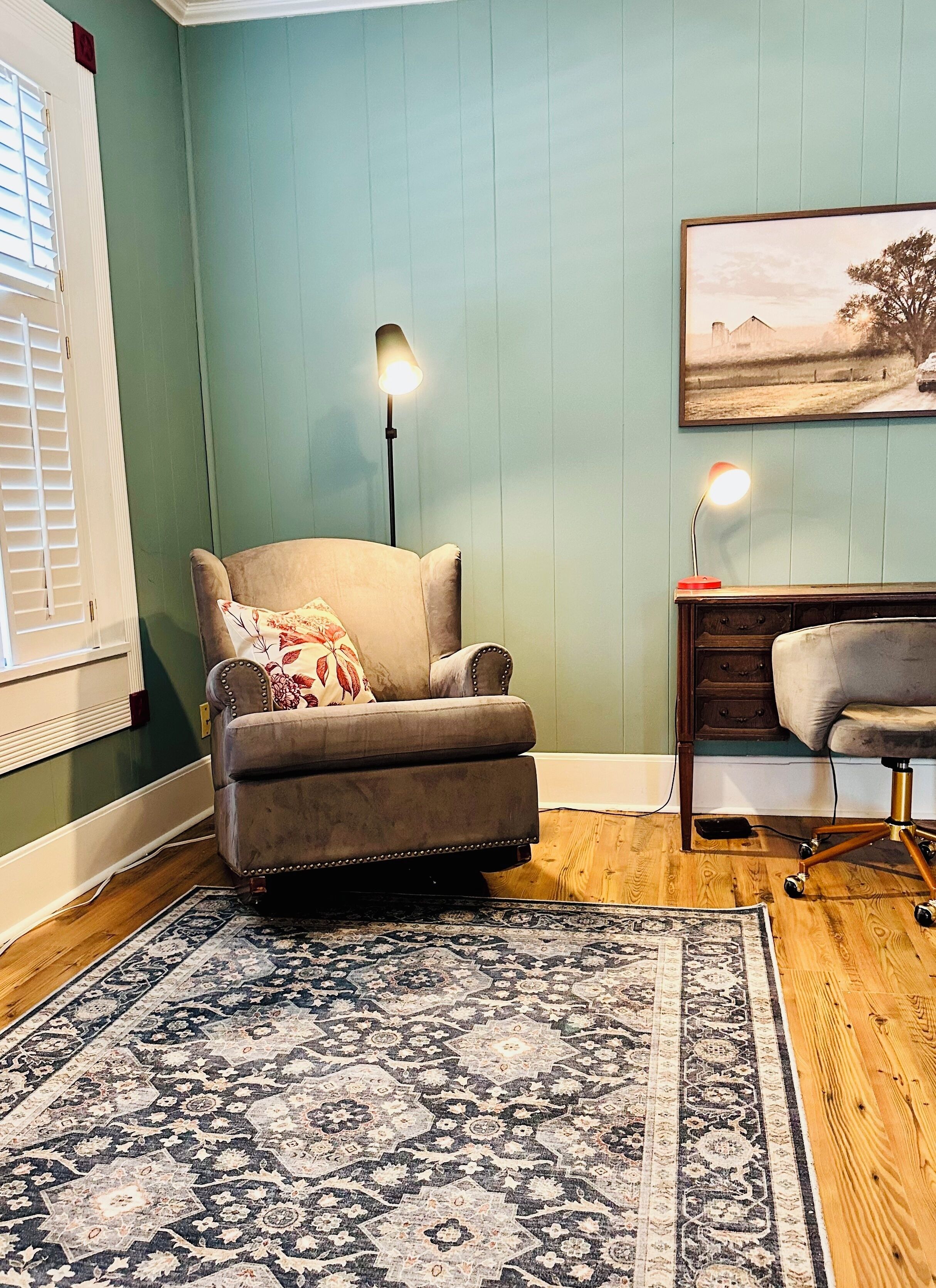 Een kamer met een grijze fauteuil, een kamerbreed tapijt met patroon, een bureau met lamp en een schilderij aan de muur.