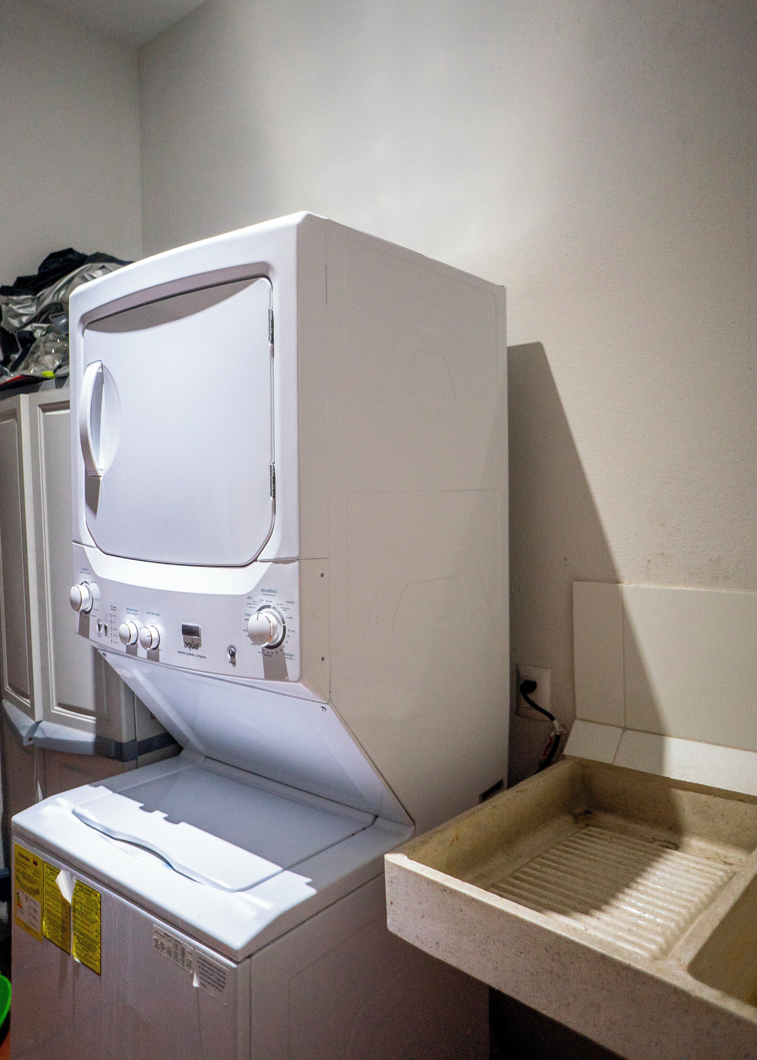 Lavadora blanca compacta con panel de control, ubicada en un pequeño cuarto de lavado con pared de azulejos y un lavamanos.