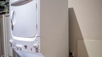 A compact, white washing machine with a control panel, situated in a small laundry room with a tiled wall and a sink.