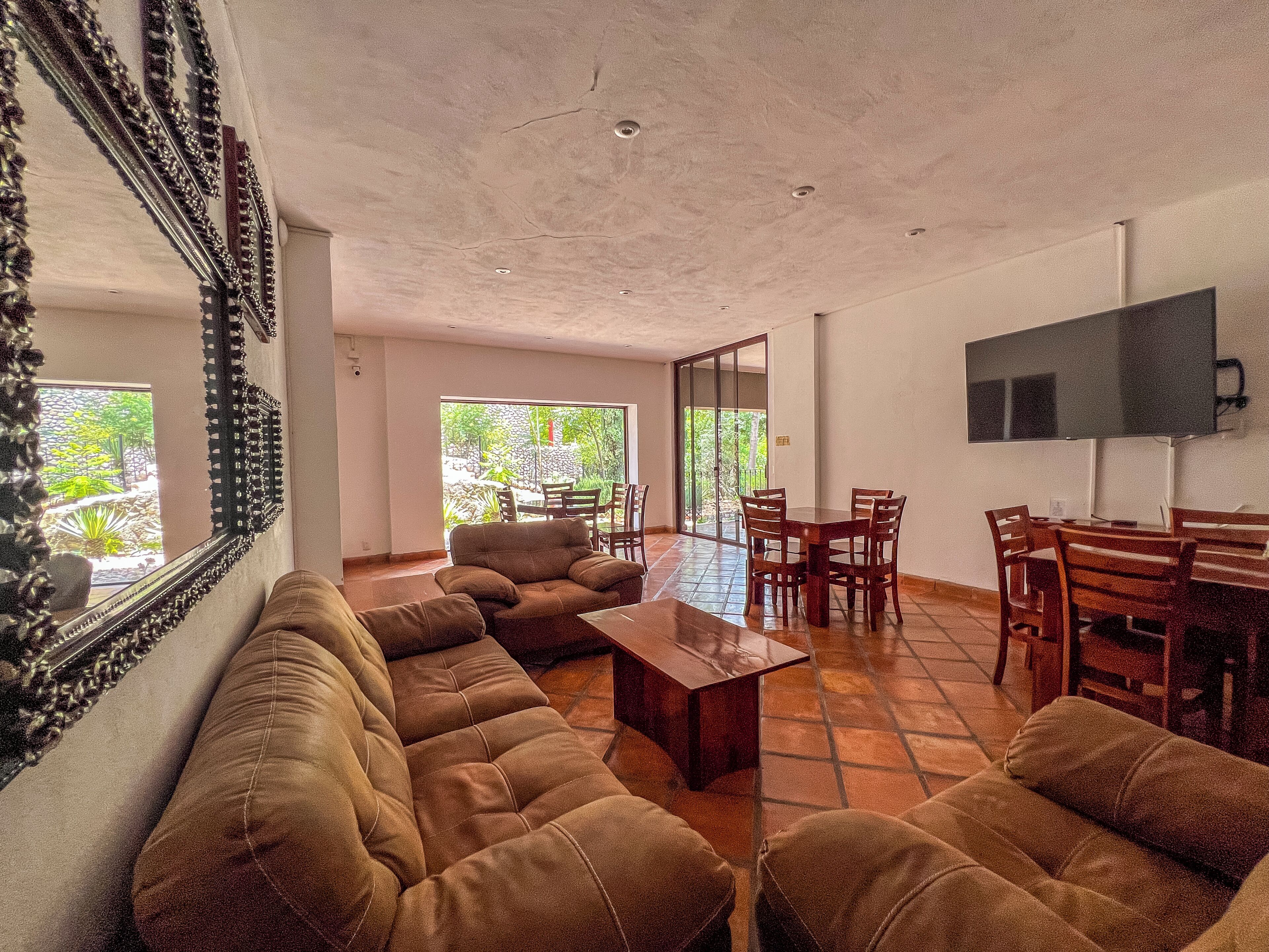 A spacious living area with a sofa set, a coffee table, and a dining table with chairs. There is a television mounted on the wall and a large window providing a view of the outdoors.