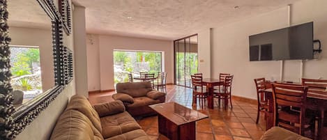 A spacious living area with a sofa set, a coffee table, and a dining table with chairs. There is a television mounted on the wall and a large window providing a view of the outdoors.