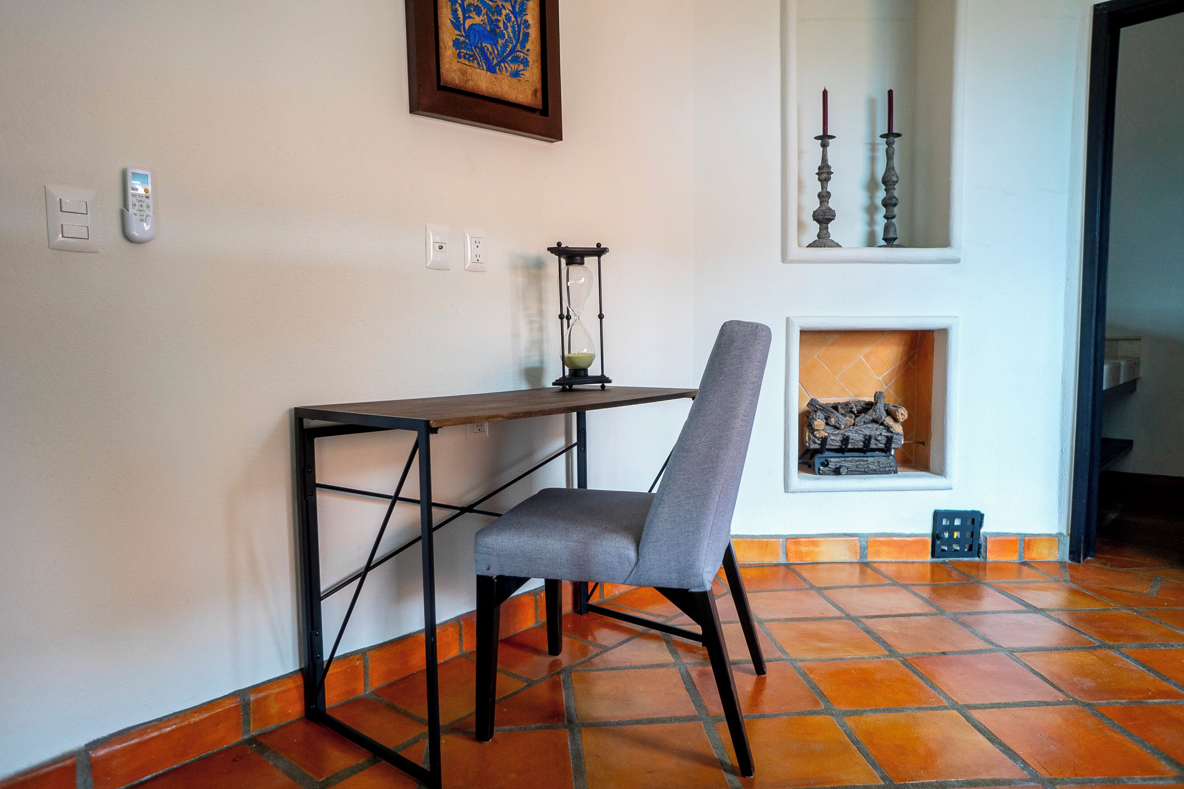 A room with a wooden desk, a grey chair, a wall-mounted hourglass, and a fireplace with a log.