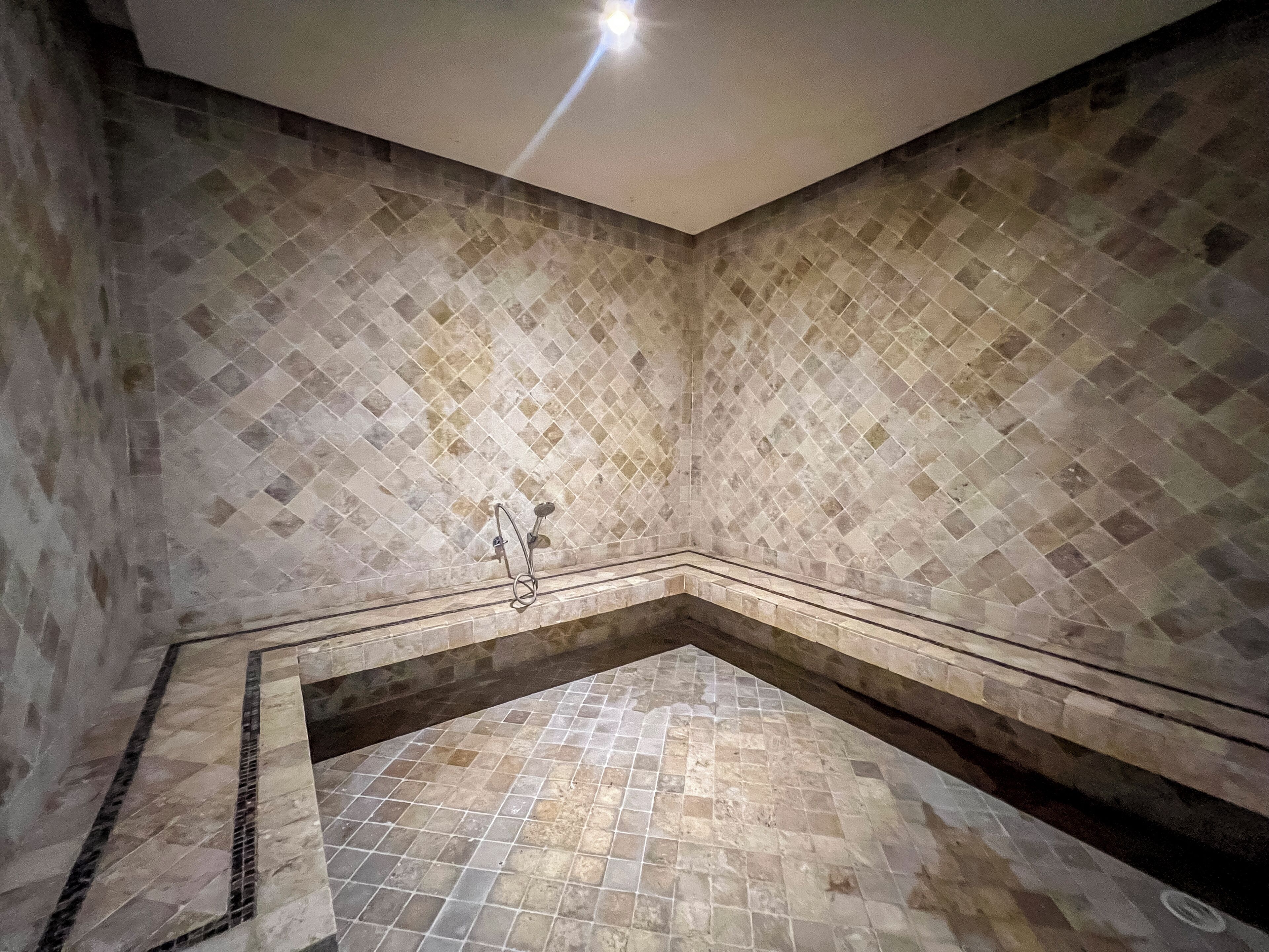 A corner bathtub with a tiled floor and walls, a showerhead, and a drain.