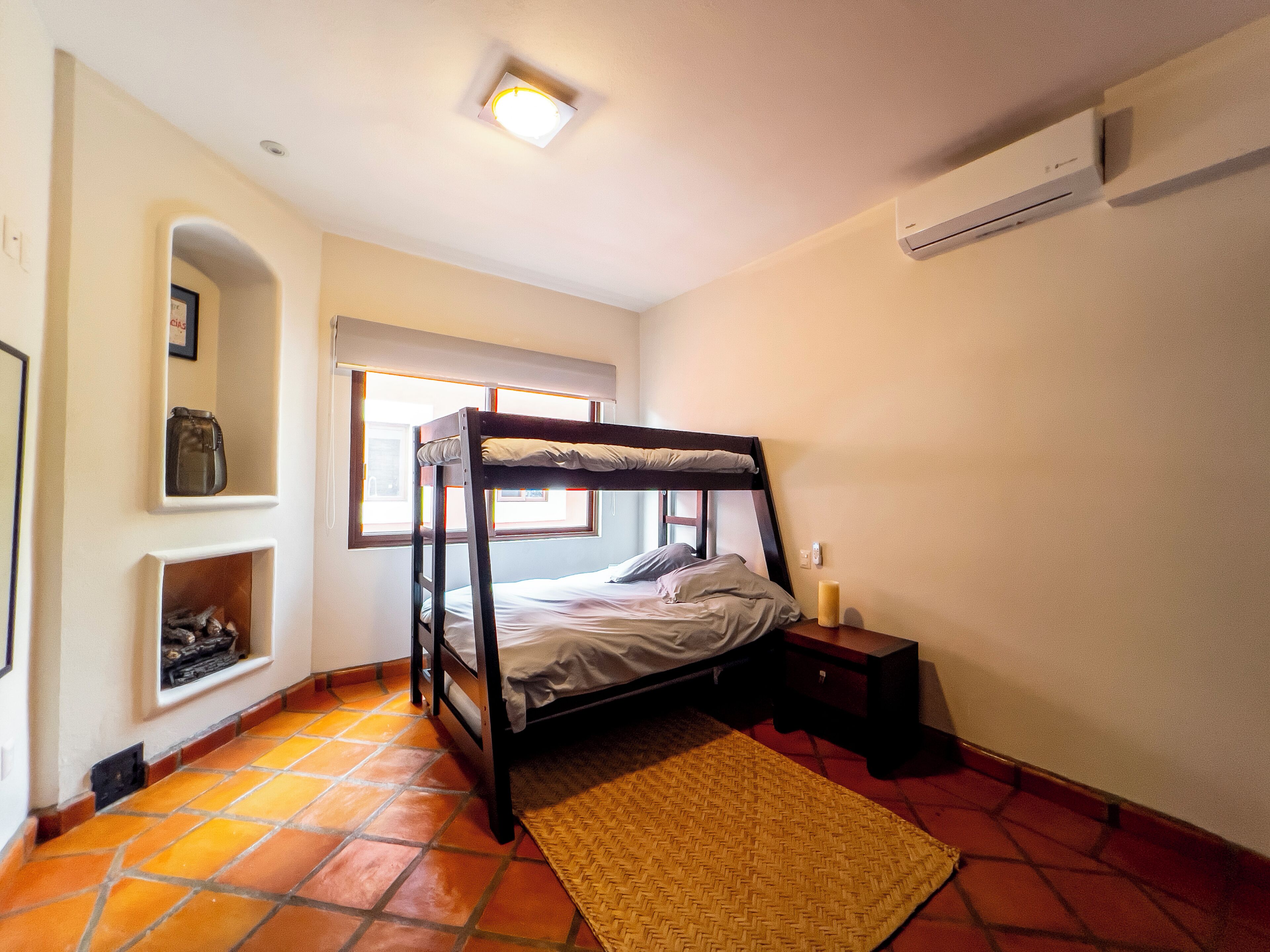 A room with a bunk bed, a small bedside table, a shelf with a framed picture, a window with blinds, and a tiled floor.