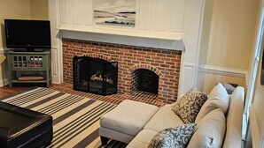 A living room with a brick fireplace, a flat-screen TV on a cabinet, a striped rug, and a light-colored sofa with patterned cushions. - 3-bedroom house in Durham near UNC, Duke and the American Tobacco Trail (Durham)