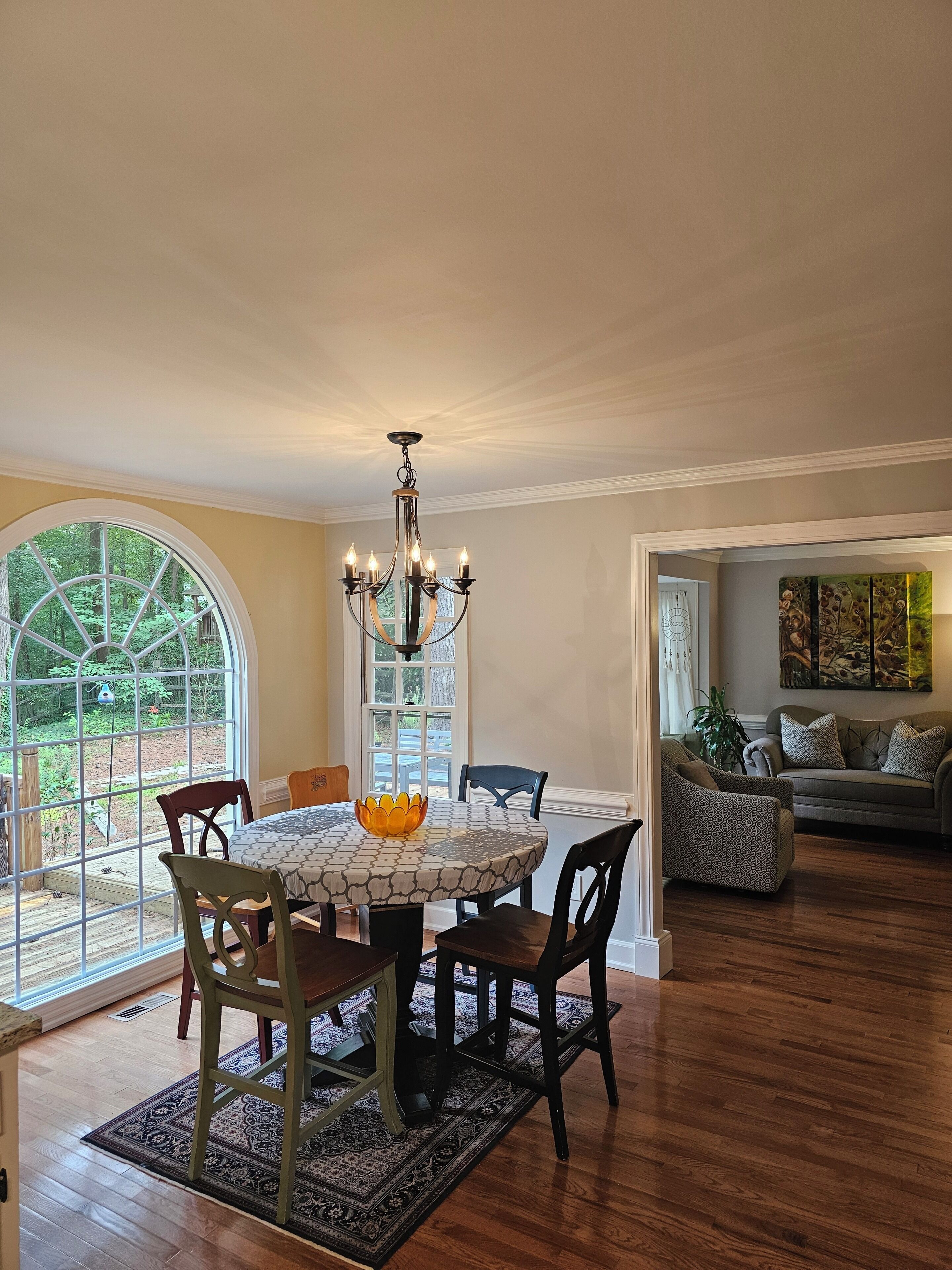 A dining area with a round table, chairs, a chandelier, and a view through a large window.