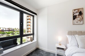 Une chambre d’hôtel avec une grande fenêtre, une table de chevet avec une lampe, un lit avec du linge de lit blanc et un tableau au mur.