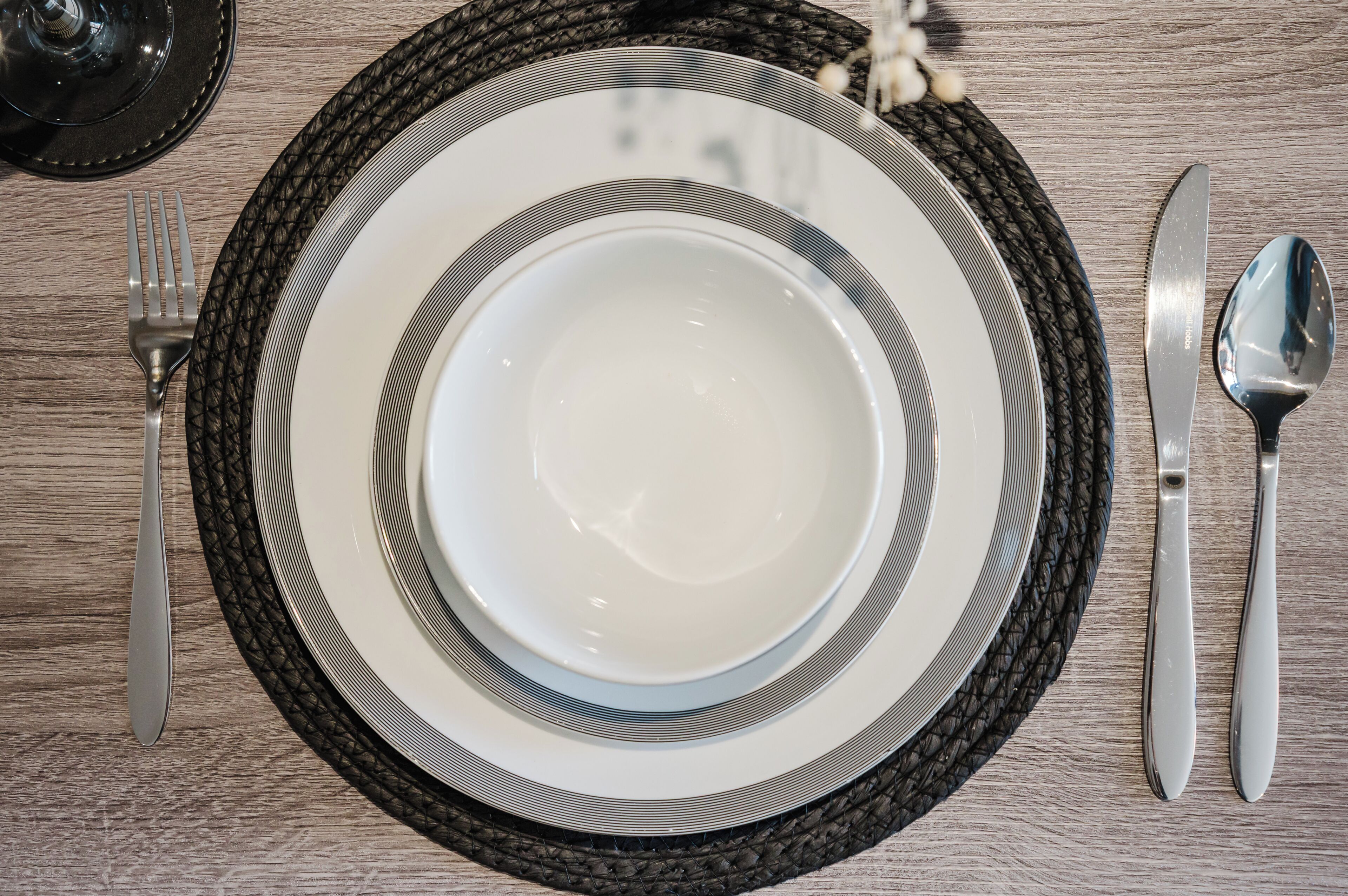 A table set with a white plate, a black rimmed bowl, a fork, a knife, and a spoon on a woven placemat.