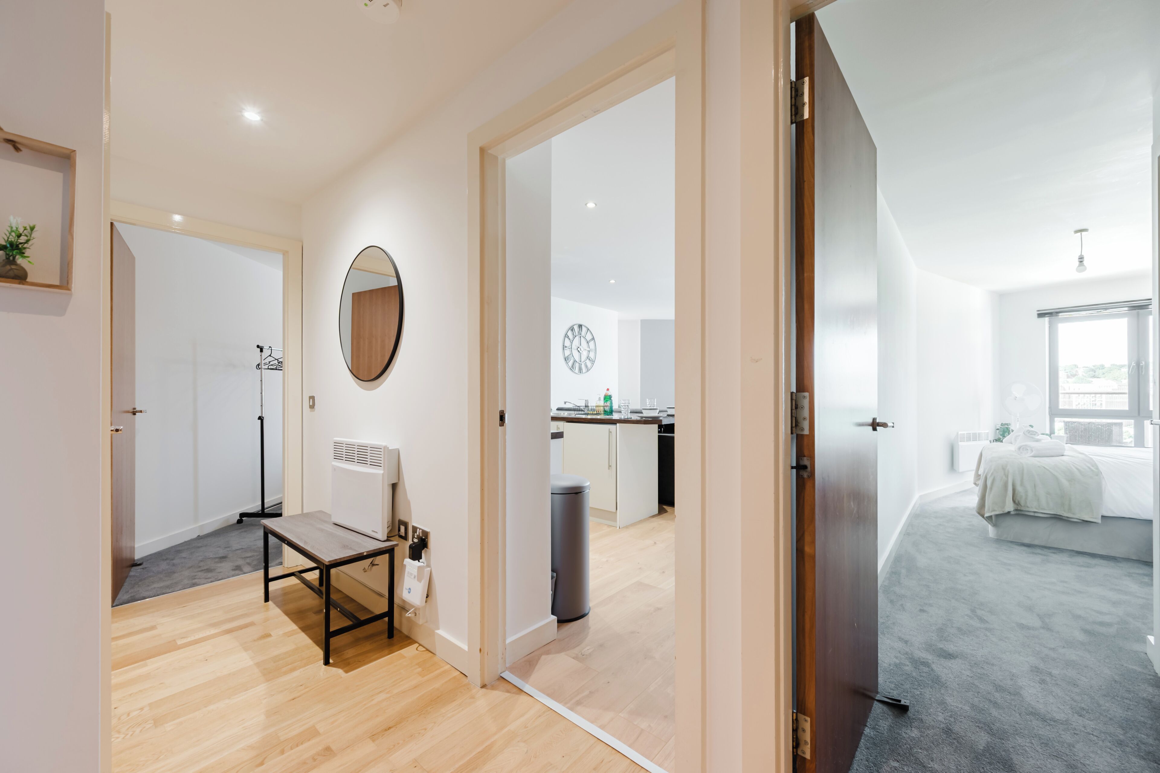 Un intérieur moderne avec un plancher en bois, un miroir, un radiateur et un coin cuisine visible à travers une porte ouverte.