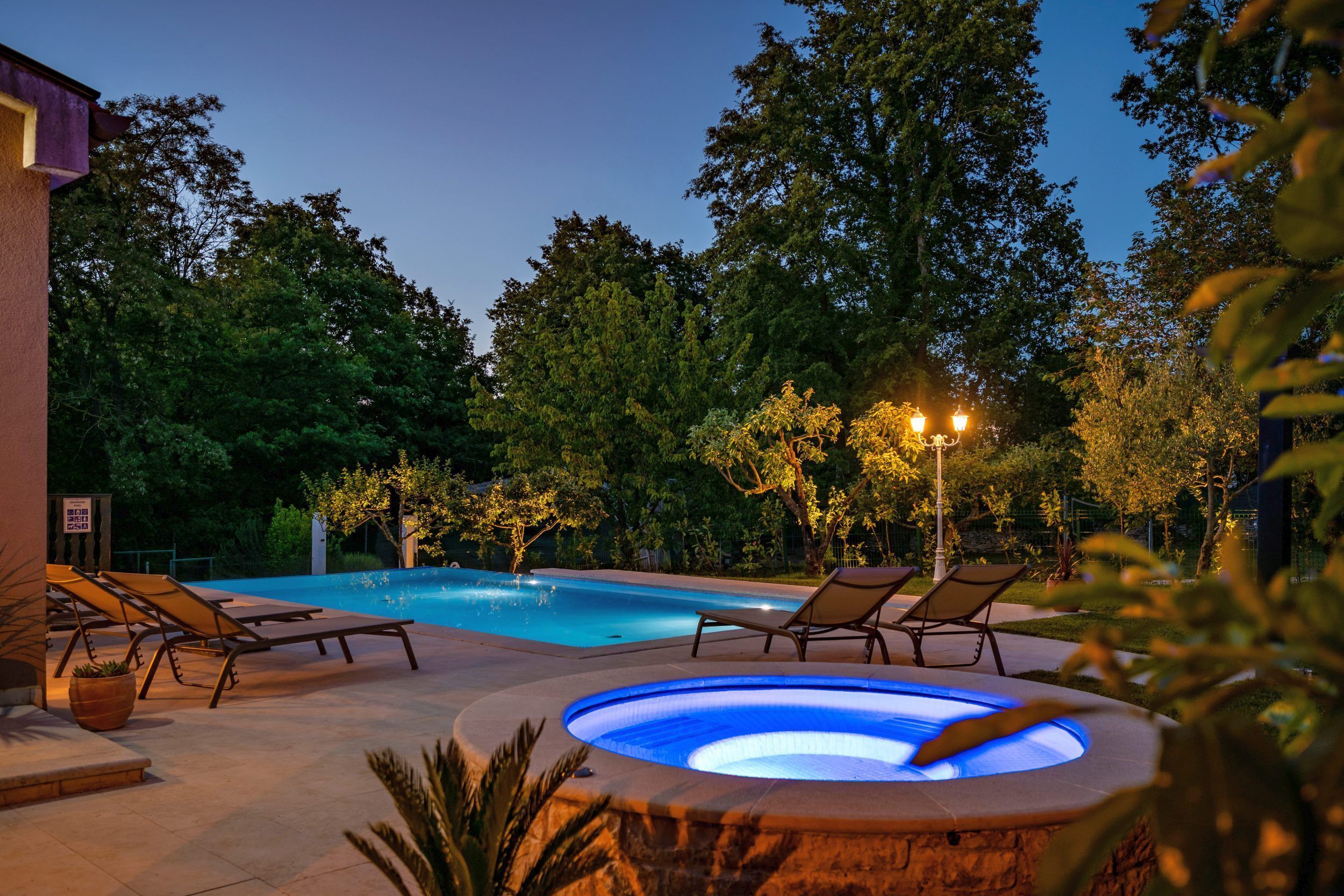 A swimming pool with lounge chairs and a hot tub, surrounded by trees and a building.