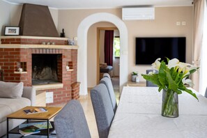 Una sala de estar con chimenea de ladrillo, un televisor de pantalla plana montado en la pared y una mesa de comedor con sillas.