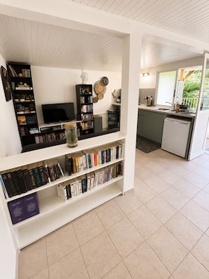 A modern living room with a flat-screen TV, a bookshelf, and a dining area with a kitchen in the background. - L'Escapade Ludique | 80 Board Games & Private Terrace (Saint-Esprit)