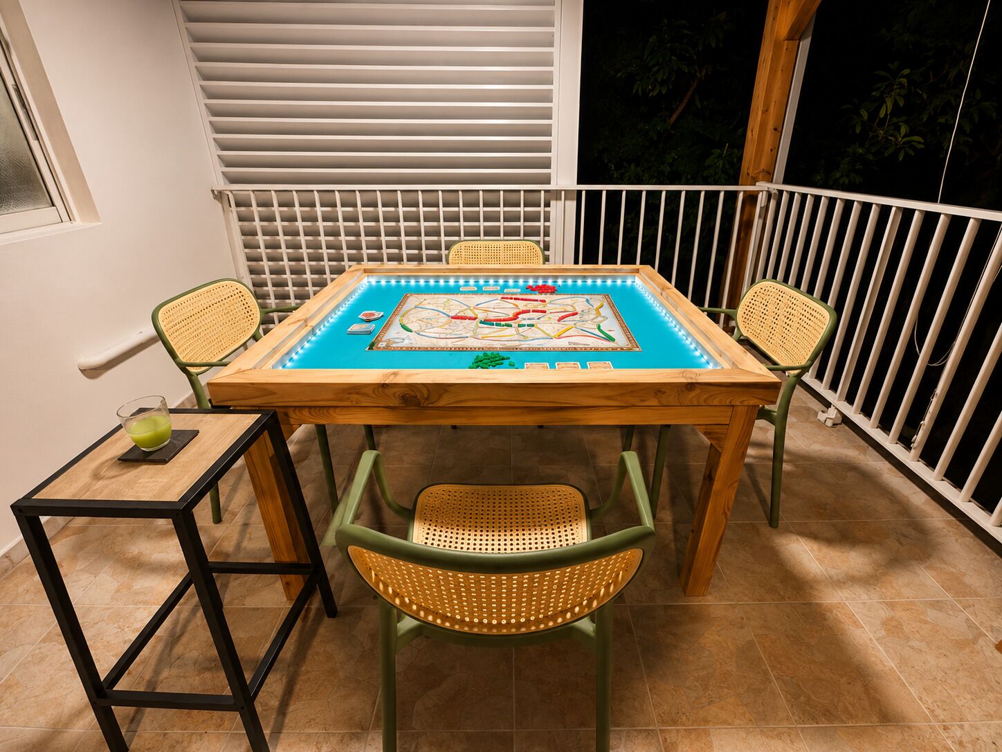 A wooden table with a board game, surrounded by wicker chairs, on a balcony with a railing.