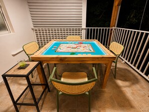 Uma mesa de madeira com um jogo de tabuleiro, rodeada por cadeiras de vime, numa varanda com guarda.