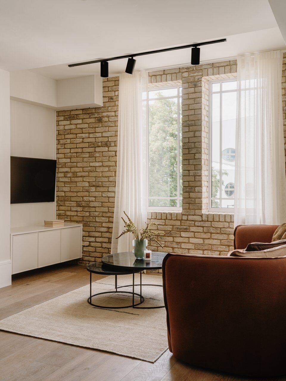 Un salon moderne avec un mur en briques, un téléviseur à écran plat fixé au mur, un canapé marron et une petite table ronde sur laquelle est posé un vase de fleurs.