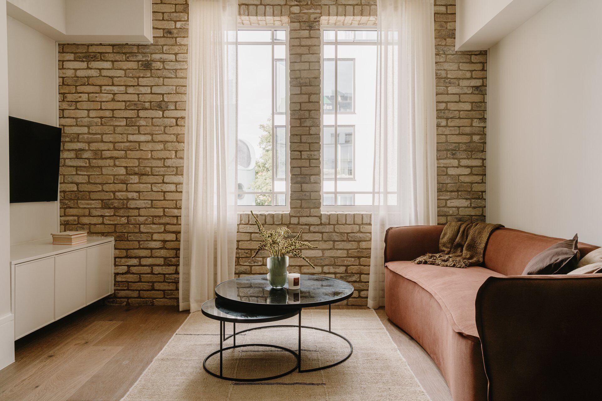 A living room with a brick wall, a round black table with a vase and candle, a brown sofa, and a TV on the wall.