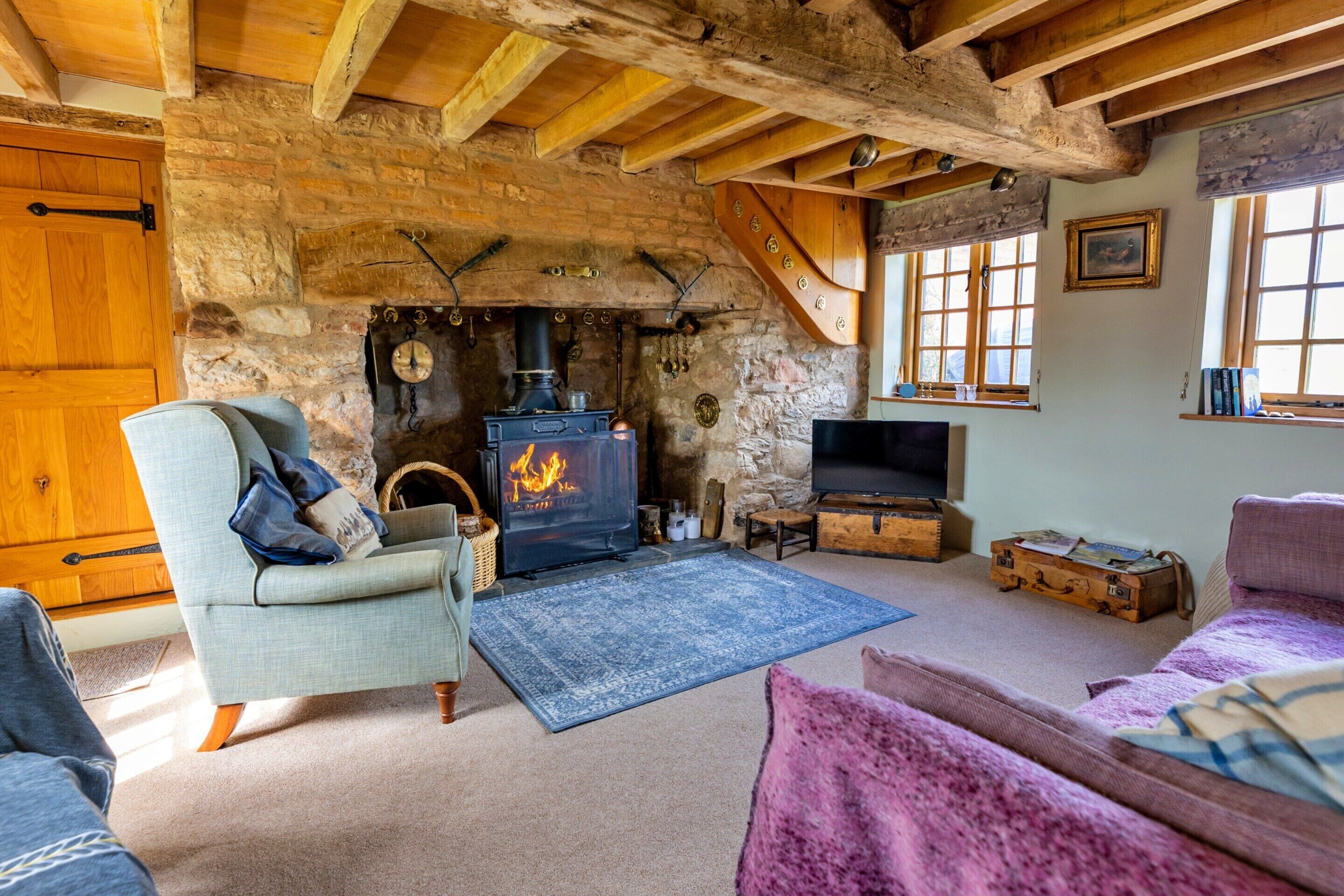 A rustic interior with exposed wooden beams, a stone wall, a fireplace with a modern wood-burning stove, a television, and cozy seating.