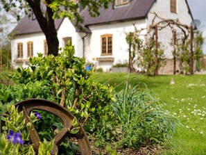 Ein Garten mit einem Metallrad, verschiedenen Pflanzen und einem weißen Gebäude mit Fenstern im Hintergrund.