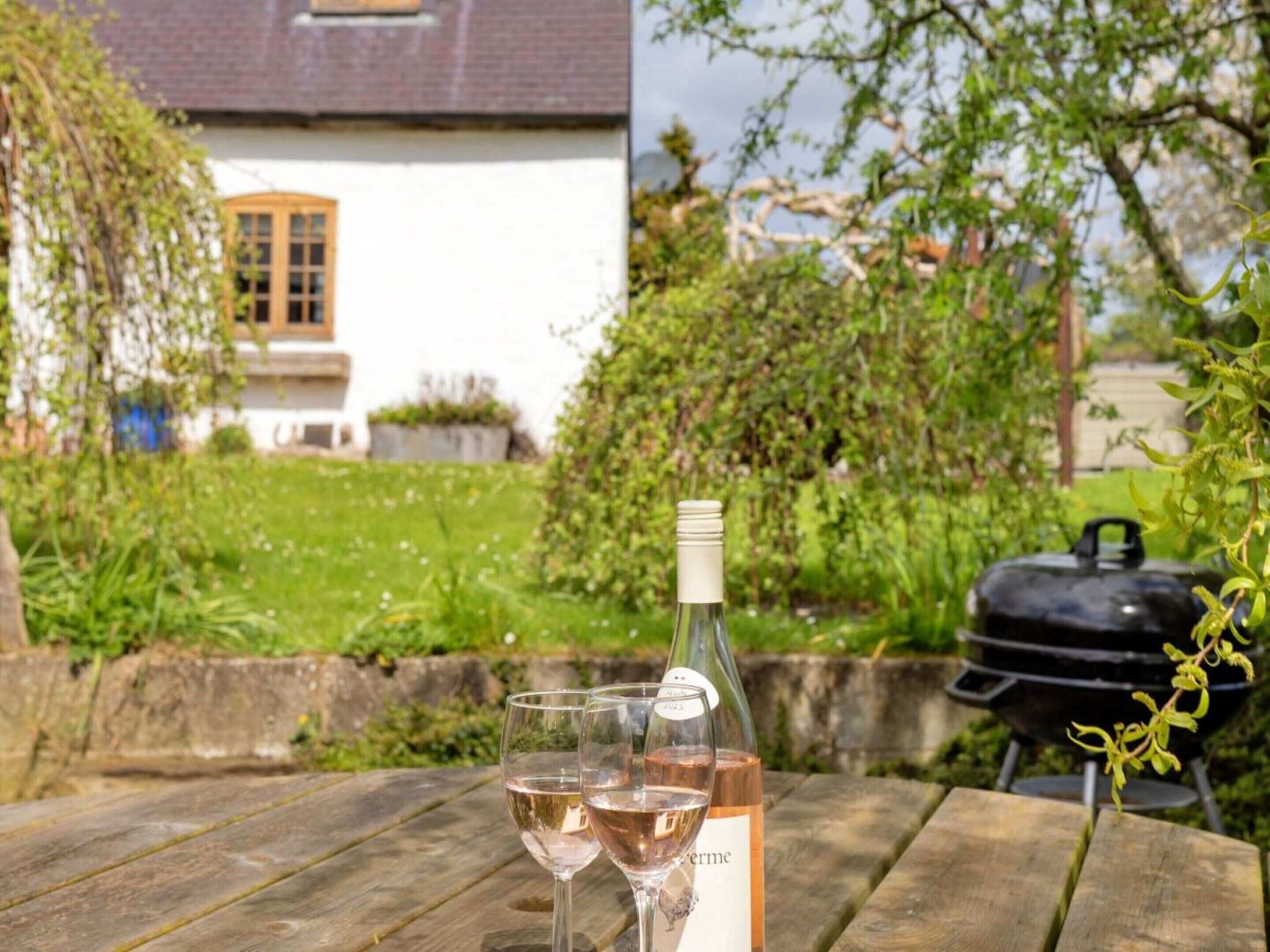 A bottle of wine and two glasses on a wooden table with a barbecue grill in the background.