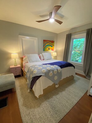 A bedroom with a bed, two bedside tables, a ceiling fan, a window with curtains, and a colorful abstract painting on the wall. - Designer 2 bedroom cottage minutes from downtown where luxury meets convenience. (Charlotte)