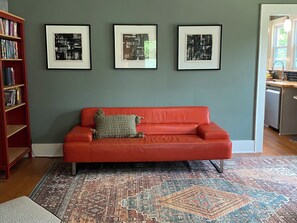 A red sofa with a patterned cushion, a large rug, and three framed abstract artworks on a wall. - Designer 2 bedroom cottage minutes from downtown where luxury meets convenience. (Charlotte)