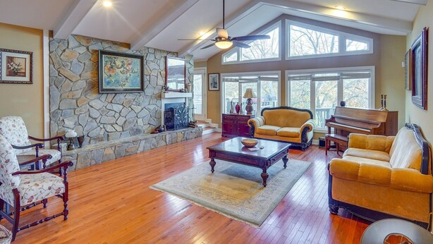 A spacious living room with a stone fireplace, large windows, and a ceiling fan.