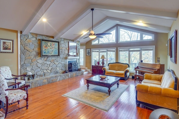 A spacious living room with a stone fireplace, large windows, and a ceiling fan.