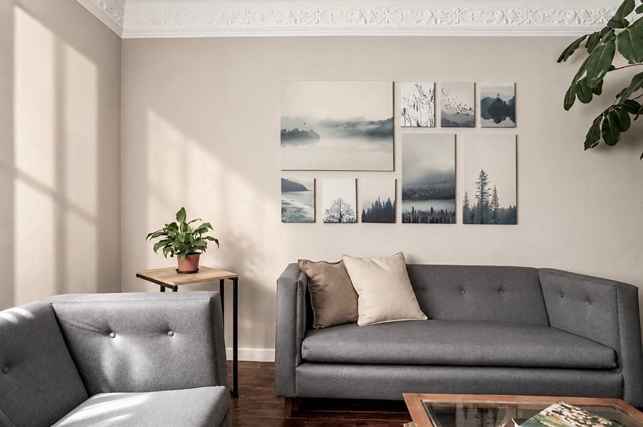 En stue med en grå sofa, en matchende lænestol, et lille bord med en potteplante og en væg med indrammede kunstværker.