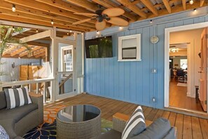 Ein überdachter Außenbereich mit Deckenventilator, einem an der Wand befestigten Fernseher und einer Sitzecke mit Sofa und rundem Tisch.