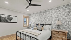Une chambre à coucher comprenant un lit, une table de chevet, une lampe, un ventilateur de plafond, une fenêtre et un mur recouvert d’un papier peint à motifs floraux.