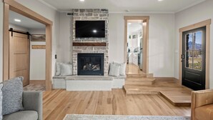 Un salon moderne avec une cheminée en pierre, un téléviseur à écran plat et une vue sur la cuisine grâce à une porte ouverte.