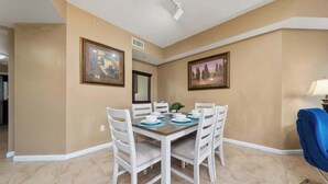 Un comedor con una mesa preparada para cuatro personas, rodeada de sillas blancas, en una habitación con paredes color crema y piso de baldosas.