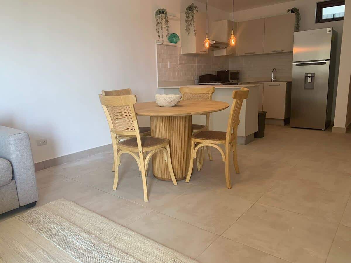 A modern kitchen with a dining area, featuring a round table and wicker chairs, a grey sofa, and a stainless steel refrigerator.